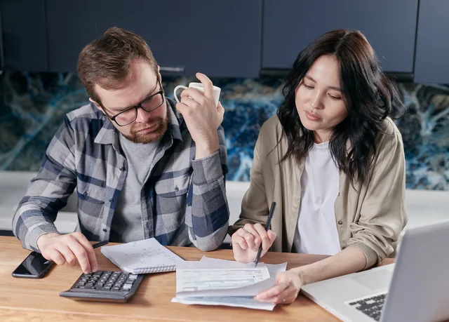A couple reviewing household bills and budget using a calculator and laptop at their kitchen table. Creator payment terms in the UK: invoice lines, VAT and late payment